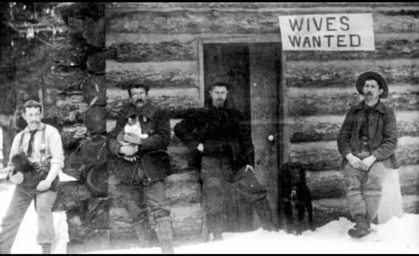 black and white photo of men outside a log cabin with a sign that says wives wanted, Funny Fake history photos, r fakehistoryporn, facts about history that are not true, false textbook photos, historical pics with funny captions, lol, jokes, old photos with hilarious explanations, funny pics